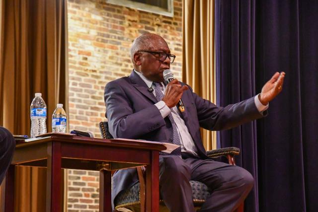 **Alt text:** Representative Jim Clyburn speaking into a microphone during a town hall event in Summerton, South Carolina, in April, seated on stage beside a small table with water bottles while gesturing to the audience.